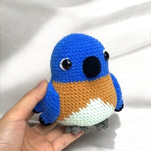 Puede incluir: Un juguete de peluche de p&aacute;jaro tejido a crochet en azul, naranja y blanco con ojos negros y un pico negro. El p&aacute;jaro tiene peque&ntilde;as patas grises.