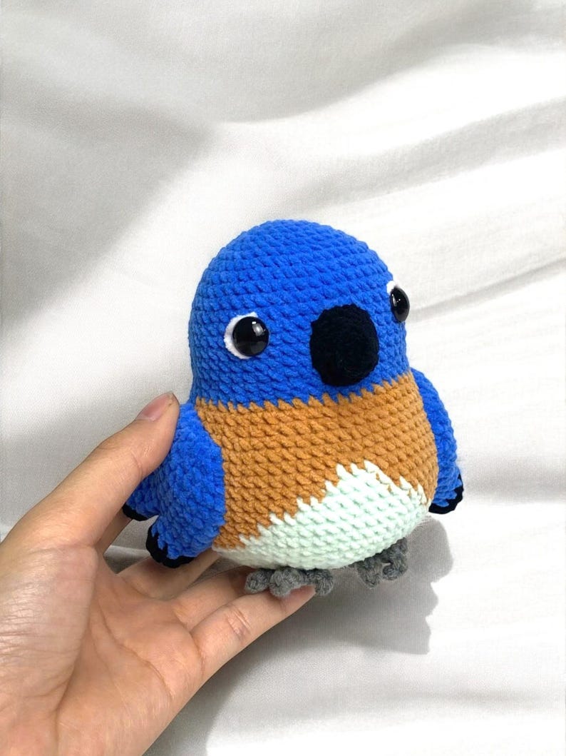 Puede incluir: Un juguete de peluche de p&aacute;jaro tejido a crochet en azul, naranja y blanco con ojos negros y un pico negro. El p&aacute;jaro tiene peque&ntilde;as patas grises.