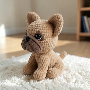 Puede incluir: Un juguete de peluche de bulldog francés tejido a ganchillo, en tonos beige y marrón. El perro tiene ojos grandes y oscuros y un hocico detallado. Está sentado sobre una superficie blanca texturizada, mostrando la artesanía.