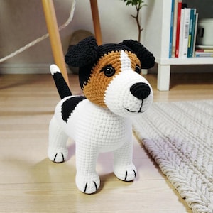May include: A crocheted Jack Russell Terrier dog toy. The toy is white with black and tan markings, black ears, and a black tail. The dog is standing on a light-colored rug. The toy is handmade.