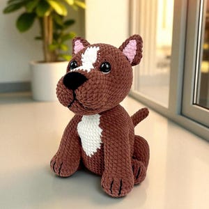 May include: A brown crochet dog with a white marking on its head and chest. The dog has pink inner ears and a black nose. The soft toy is sitting, with its tail up. A handmade item.
