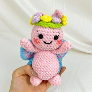 May include: A handmade pink crochet butterfly doll with a flower crown. The doll has blue and pink wings, rosy cheeks, and a smiling face. The flower crown features yellow, purple, and white flowers with a green base.