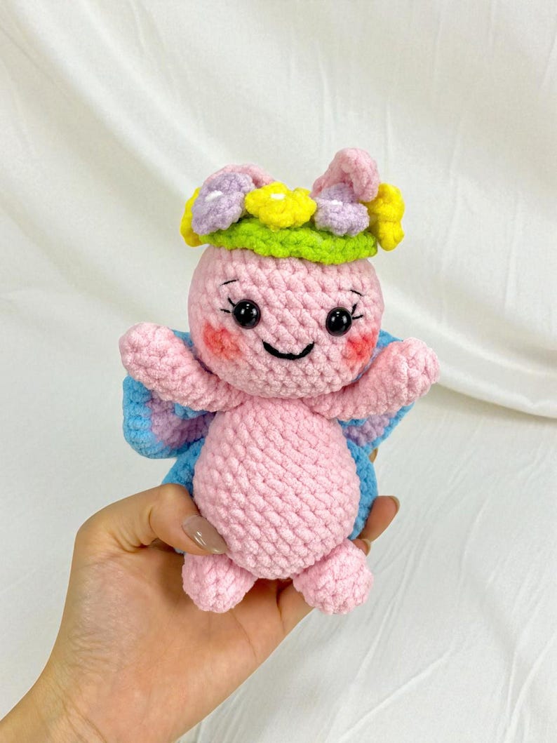 May include: A handmade pink crochet butterfly doll with a flower crown. The doll has blue and pink wings, rosy cheeks, and a smiling face. The flower crown features yellow, purple, and white flowers with a green base.