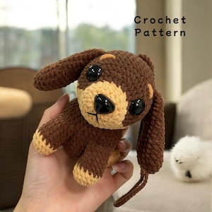 May include: Crochet pattern for a brown and tan crocheted dog with black eyes. The dog is sitting on a hand and has a long tail.