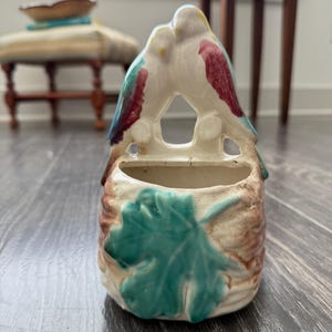 Könnte beinhalten: Ein Vintage-Wandtaschen-Pflanzgefäß aus Keramik mit zwei Vögeln, die zusammen sitzen. Die Vögel haben gelbe Köpfe, rote Flügel und blaue Akzente. Der Pflanztopf hat eine braune und cremefarbene Basis mit einem grünen Blattdesign.