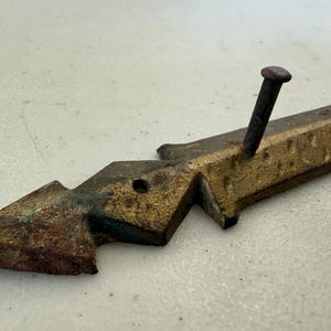 May include: A weathered, antique metal architectural element with a nail. The piece has a gold and brown finish with a geometric design. The nail is dark brown and is inserted into the metal.