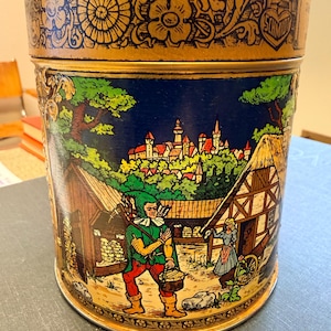 Könnte beinhalten: Eine zylindrische Metalldose mit einem goldfarbenen Deckel, der mit geprägten Blumen- und Rankenmustern verziert ist. Der Körper zeigt eine farbenfrohe Illustration einer mittelalterlichen Dorfszene mit einer Person in grüner Tunika und roter Hose.
