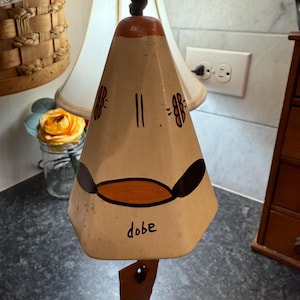 Può includere: Una campana color crema con la parte superiore marrone e un battaglio in pelle. La campana presenta dettagli dipinti in nero e arancione, inclusa la parola "dobe". È appesa a un cordoncino marrone.