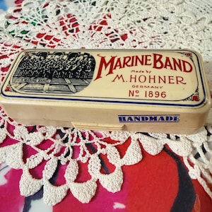 Puede incluir: Un estuche rectangular de color crema para armónica Marine Band. El estuche presenta el texto "MARINE BAND" en escritura roja, "Made by M. Hohner Germany No. 1896" y "HANDMADE" en azul. Una ilustración de una banda está en el lado izquierdo.