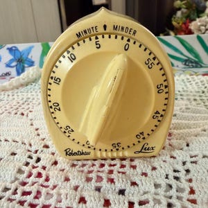 May include: A vintage, cream-colored kitchen timer with the words "MINUTE MINDER" at the top. The timer has a large dial with numbers and a prominent knob. The brand names "Robertshaw" and "Lux" are also visible.
