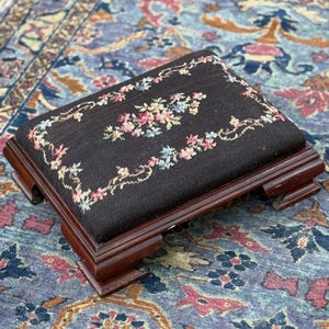 Puede incluir: Un pequeño reposapiés rectangular con una base de madera marrón oscuro y una parte superior de tela negra. La parte superior está bordada con un patrón floral en tonos de rosa, azul y amarillo. El reposapiés está colocado sobre una alfombra estampada.