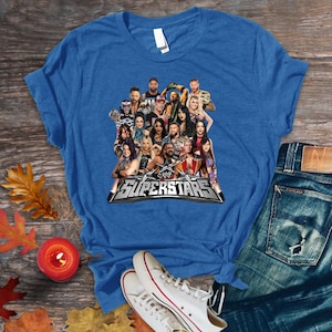 May include: Blue t-shirt featuring a graphic of multiple WWE Superstars. The graphic includes the text "WWE Superstars" in silver and black. The shirt is displayed on a wooden surface with jeans and sneakers.