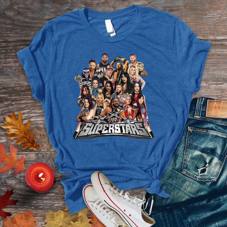 May include: Blue t-shirt featuring a graphic of multiple WWE Superstars. The graphic includes the text "WWE Superstars" in silver and black. The shirt is displayed on a wooden surface with jeans and sneakers.