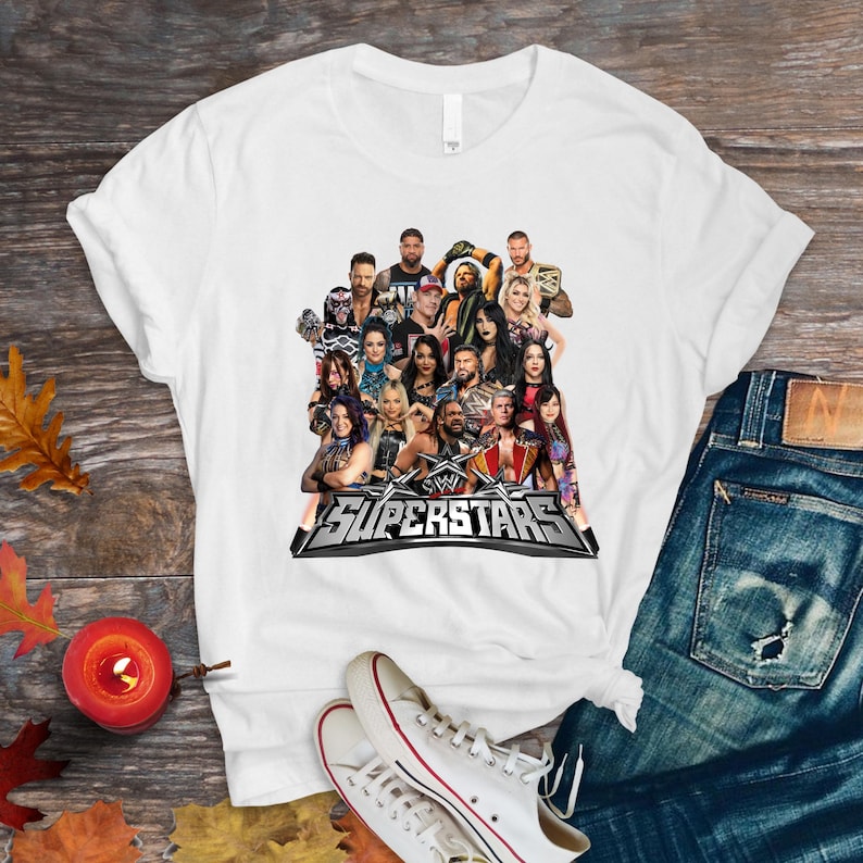 May include: White t-shirt featuring a graphic of multiple WWE Superstars. The design includes the WWE logo and the word "Superstars" in a silver and black design. The shirt is displayed on a wooden surface with jeans and sneakers.