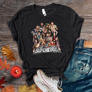 May include: Dark gray t-shirt featuring a graphic of multiple WWE Superstars. The word "Superstars" is displayed in a silver and black design. The shirt is paired with blue jeans and white sneakers.