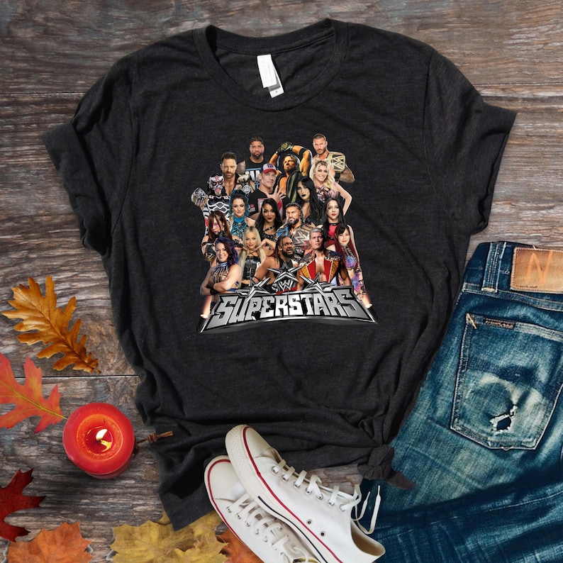 May include: Dark gray t-shirt featuring a graphic of multiple WWE Superstars. The word "Superstars" is displayed in a silver and black design. The shirt is paired with blue jeans and white sneakers.