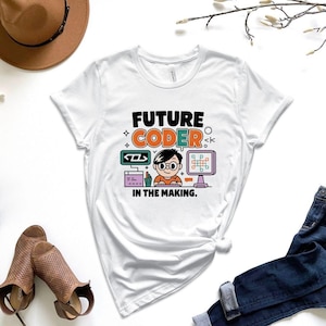 May include: White t-shirt with the text "FUTURE CODER IN THE MAKING" and a colorful graphic of a person with glasses in front of two computer monitors. The shirt is paired with blue jeans and brown shoes.