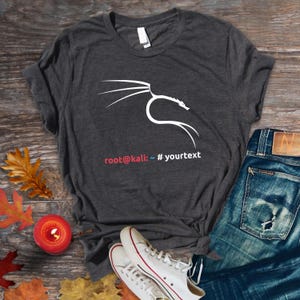 May include: Dark gray t-shirt featuring a white dragon graphic and the text "root@kali: ~ # yourtext". The shirt is paired with blue jeans and white sneakers with red accents. A candle and autumn leaves are in the background.