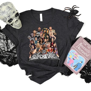 May include: Dark gray t-shirt featuring a graphic of multiple WWE Superstars and the text "WWE SUPERSTARS" in a silver and red design. The shirt is displayed with Halloween decorations, including a skull, spider, and a book.