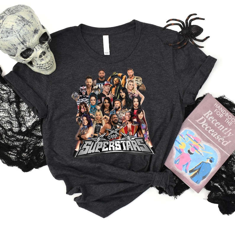 May include: Dark gray t-shirt featuring a graphic of multiple WWE Superstars and the text "WWE SUPERSTARS" in a silver and red design. The shirt is displayed with Halloween decorations, including a skull, spider, and a book.
