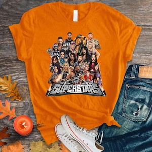 May include: An orange t-shirt featuring a group of WWE Superstars. The shirt displays a collage of wrestlers with the text "WWE Superstars" in a silver and black design. The shirt is paired with blue jeans and white sneakers.