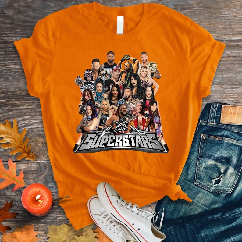 May include: An orange t-shirt featuring a group of WWE Superstars. The shirt displays a collage of wrestlers with the text "WWE Superstars" in a silver and black design. The shirt is paired with blue jeans and white sneakers.