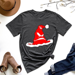 May include: Dark gray t-shirt featuring a red Santa hat graphic with a white trim and a white pom-pom. The graphic has a stylized design. The shirt is paired with blue jeans and brown shoes.