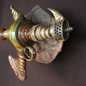 Può includere: Una scultura in ceramica di un'astronave stilizzata con accenti verdi, marroni e dorati. L'astronave ha un corpo lungo e sottile con un naso appuntito e un grande motore rotondo sul retro. La scultura è montata su un pezzo di ceramica rotta.