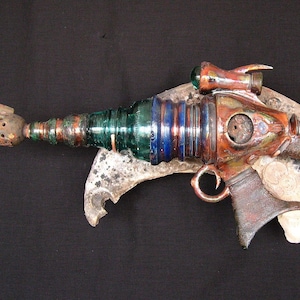 Può includere: Una pistola di stile steampunk, realizzata a mano, realizzata con materiali riciclati. La pistola è realizzata in metallo, vetro e legno e presenta una canna in vetro verde, dettagli in rame e un calcio in legno marrone.