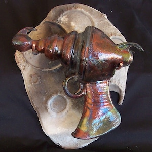 Può includere: Una scultura in ceramica di una pistola a raggi futuristica con una finitura marrone metallizzata e iridescente. La pistola è montata su una base in ceramica grigia testurizzata.