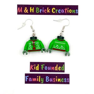 May include: A pair of green Lego-style earrings with a Christmas sweater design. The earrings have silver hooks and gray hands. The image includes the text "M & H Brick Creations" and "Kid Founded Family Business".