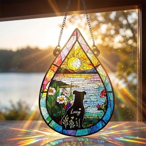 May include: A teardrop-shaped stained glass suncatcher featuring a landscape scene with a yellow sun, blue water, and white daisies. A black silhouette of a dog is centered with the name Lucy and a small heart printed in white script.