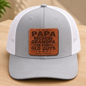 May include: A light gray and white trucker hat featuring a square brown leather patch on the front. The patch is engraved with the text "PAPA BECAUSE GRANDPA IS FOR OLD GUYS" and the names "Sophia • Thomas" at the bottom with decorative stars.