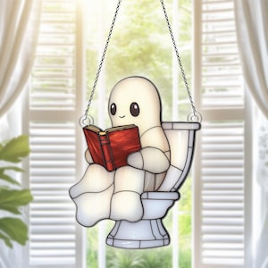 May include: A stained glass window hanging features a white ghost sitting on a toilet and reading a red book. The ghost has a happy expression on its face.