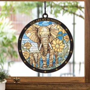 May include: A stained glass suncatcher with a round shape and a black metal frame. The suncatcher features a mother elephant and baby elephant standing in a field of sunflowers. The text "You are my sunshine" is written in a black font around the edge of the suncatcher.