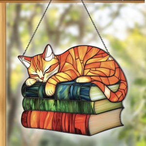 May include: A stained glass window hanging with a depiction of an orange tabby cat sleeping on top of a stack of books. The cat is curled up with its head resting on the top book. The books are stacked in a pile of three, with the top book being the largest.