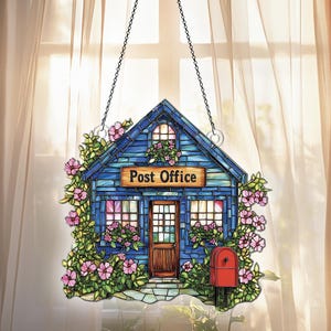May include: A stained glass window hanging with a blue post office building with a red mailbox and pink flowers. The sign above the door reads "Post Office".