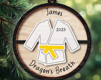 May include: A wooden ornament featuring a white karate uniform with a yellow belt, the name "James", the year "2025", and the text "Dragon's Breath". The ornament has a black border and is hanging from a chain.