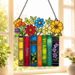 Könnte beinhalten: Ein Buntglas-Suncatcher mit einer Reihe von sechs bunten Büchern, die von leuchtenden Blumen gekrönt werden. Die Buchrücken sind gelb, rot, blau, orange, grün und dunkelblau, mit dem Text Emily, Beautiful Life und Wonderland bedruckt.