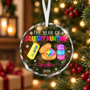 以下が含まれることがあります： THE YEAR OF SQUISHY HUNTINGのカラフルな文字、黄色いバターの塊、竹製の蒸し器に入った虹色のスクイーズ、積み重なった3つのキューブ、Sophiaという名前、2026年が描かれた、丸い透明なファセットガラスのクリスマスオーナメント。
