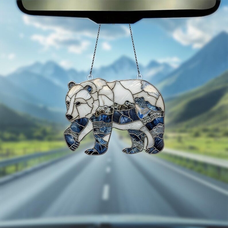 May include: A stained glass bear car charm with a white and blue design. The bear is hanging from a rearview mirror in a car. The background is a blurry image of a road and mountains.