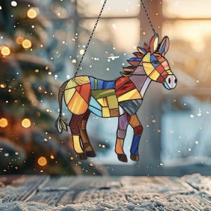Puede incluir: Un burro de vidrio coloreado con un dise&ntilde;o de patchwork. El burro est&aacute; colgado de una cadena y se encuentra frente a un fondo borroso de una ventana y un &aacute;rbol de Navidad.