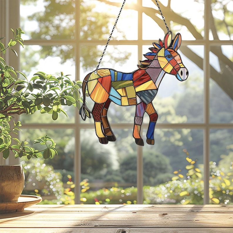 Puede incluir: Un burro de vidrio coloreado con un dise&ntilde;o de patchwork. El burro est&aacute; colgado de una cadena frente a una ventana.