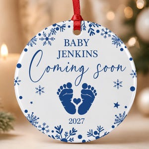 Könnte beinhalten: Ein rundes, weißes Keramikornament mit rotem Bandaufhänger, das den blauen Schriftzug BABY JENKINS Coming soon und das Jahr 2027 zeigt. Das Design enthält blaue Babyfußabdrücke mit einem kleinen Herz in der Mitte sowie blaue Schneeflocken.