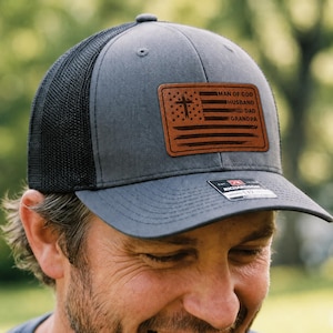 May include: A charcoal gray baseball cap with a black mesh back and a rectangular brown leather patch on the front. The patch features a laser-engraved American flag design with a cross and the text MAN OF GOD, HUSBAND, DAD, GRANDPA.