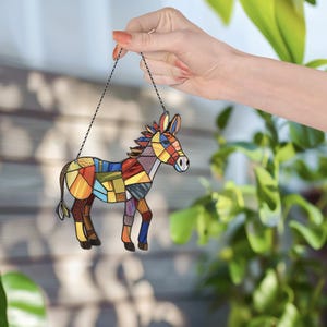 Puede incluir: Un adorno de burro de vidrio coloreado con un dise&ntilde;o de patchwork. El burro est&aacute; colgado de una cadena plateada.