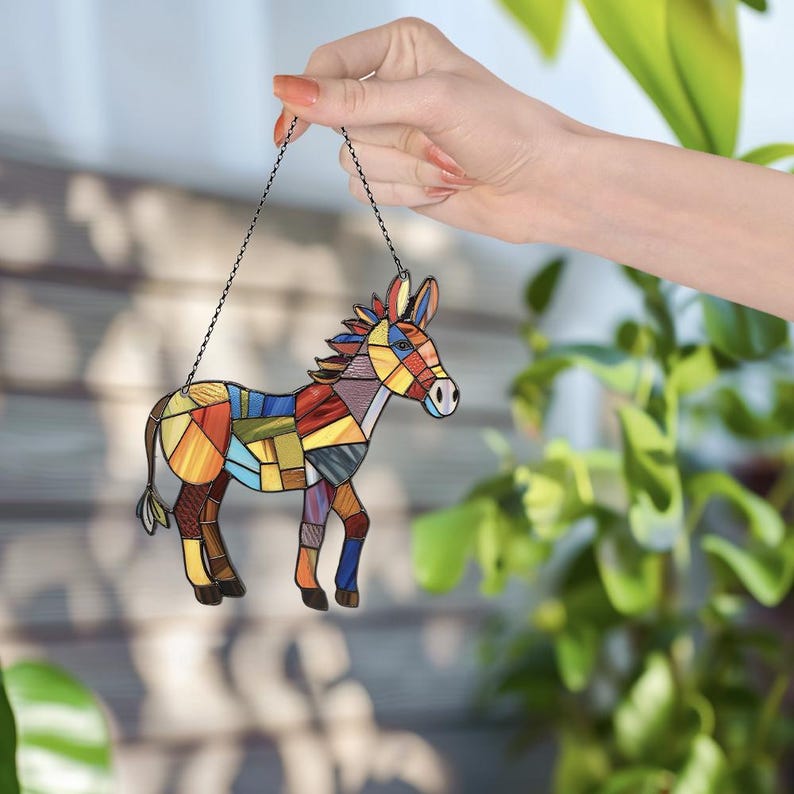 Puede incluir: Un adorno de burro de vidrio coloreado con un dise&ntilde;o de patchwork. El burro est&aacute; colgado de una cadena plateada.