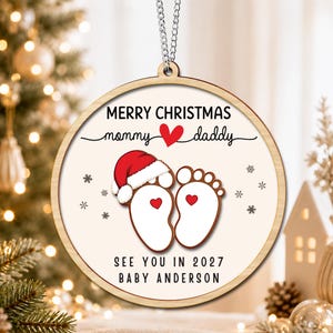 May include: A round wooden Christmas ornament featuring the text MERRY CHRISTMAS, mommy heart daddy, and SEE YOU IN 2027 BABY ANDERSON. The design includes a pair of baby footprints, one wearing a red and white Santa hat, with small red hearts.