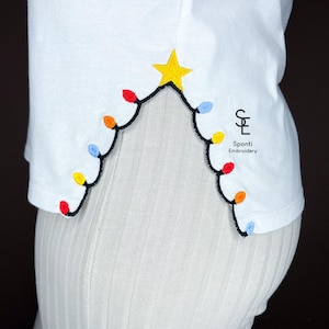 May include: White t-shirt with a Christmas light design. The design features a yellow star at the top, with a black outline of lights in red, orange, yellow, and blue. The text "Spondi Embroidery" is visible.