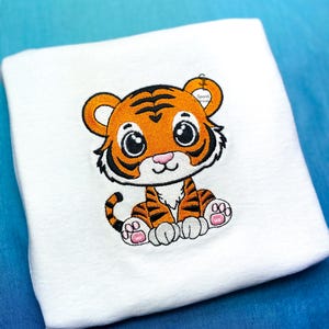 Pode incluir: Cobertor de bebê branco com um tigre de desenho animado bordado. O tigre tem listras laranja e pretas, olhos grandes e almofadas rosas. O cobertor tem uma etiqueta que diz "Sponti World".